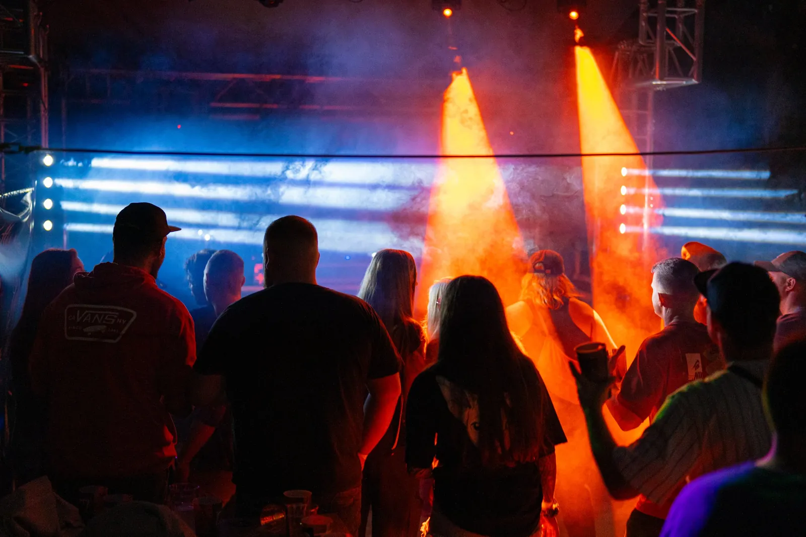 Publikum von hinten beim RAW Festival 2023, zwei orange Scheinwerferkegel schneiden durch den Bühnennebel, dahinter weiße Lichtstrahlen